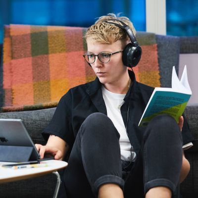 Woman with headphones sitting on floor watching something on iPad whilst holding a book